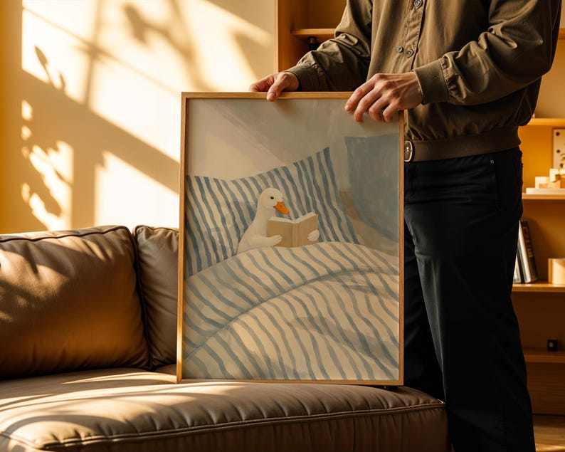 May include: A framed art print featuring a whimsical illustration of a white duck reading a book in bed. The artwork has a soft color palette with blue and beige tones, and a wooden frame.