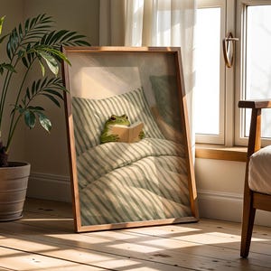 May include: A framed print featuring a whimsical illustration of a frog reading in bed. The frog is nestled under striped bedding, holding a book. The frame is a warm brown, and the scene is set in a sunlit room with a potted plant.