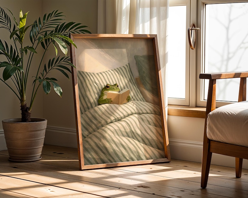 May include: A framed print featuring a whimsical illustration of a frog reading in bed. The frog is nestled under striped bedding, holding a book. The frame is a warm brown, and the scene is set in a sunlit room with a potted plant.