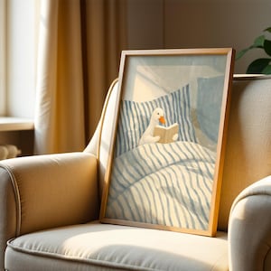 May include: A framed art print featuring a whimsical illustration of a white duck reading a book in bed. The artwork has a light blue and white striped bedding and pillow design. The frame is made of wood and is displayed on a beige armchair.