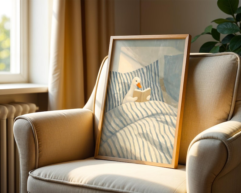 May include: A framed art print featuring a whimsical illustration of a white duck reading a book in bed. The artwork has a light blue and white striped bedding and pillow design. The frame is made of wood and is displayed on a beige armchair.
