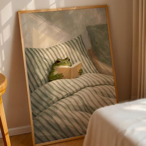 May include: A framed art print depicts a green frog reading a book in bed. The frog is nestled among striped pillows and a comforter. The artwork has a soft, warm color palette and a whimsical theme.