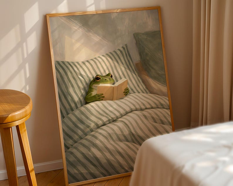 May include: A framed art print depicts a green frog reading a book in bed. The frog is nestled among striped pillows and a comforter. The artwork has a soft, warm color palette and a whimsical theme.