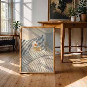 May include: A framed art print depicts a white duck reading a book in bed. The artwork features a duck with an orange beak and a beige book. The bedding has blue and white stripes. The frame is made of wood.