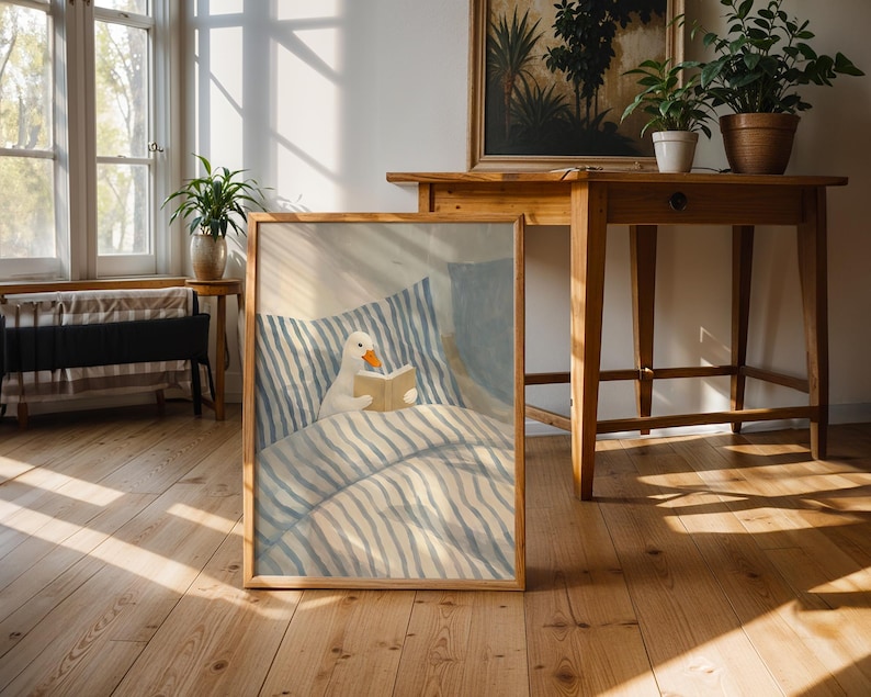 May include: A framed art print depicts a white duck reading a book in bed. The artwork features a duck with an orange beak and a beige book. The bedding has blue and white stripes. The frame is made of wood.