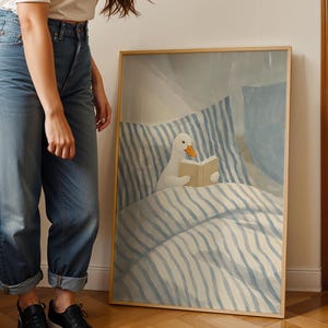 May include: A framed art print featuring a whimsical illustration of a white duck reading a book in bed. The artwork has a soft color palette of blue and beige, with striped bedding and a light wooden frame. The duck has an orange beak.