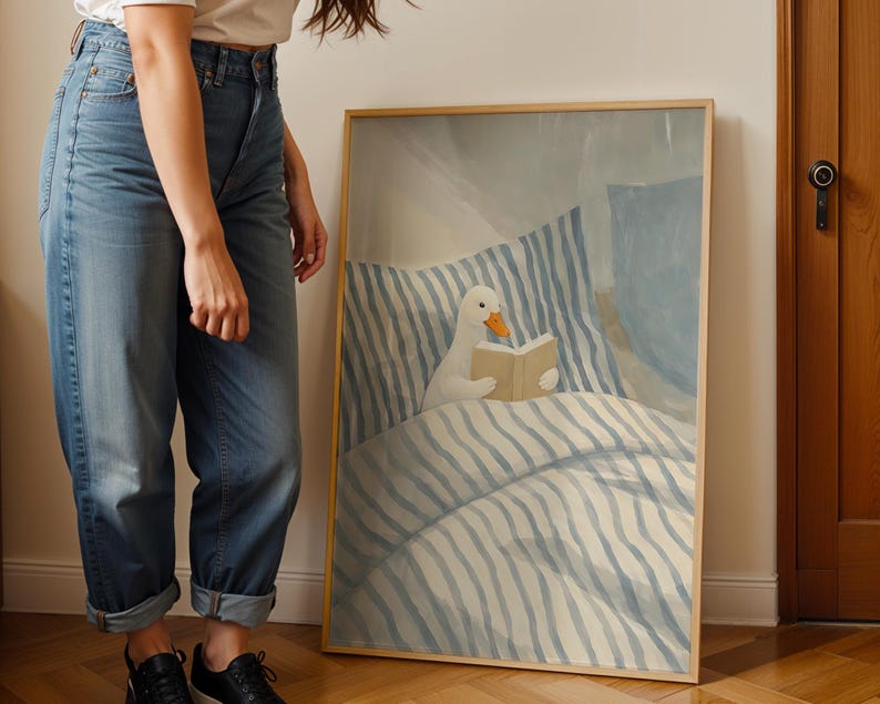 May include: A framed art print featuring a whimsical illustration of a white duck reading a book in bed. The artwork has a soft color palette of blue and beige, with striped bedding and a light wooden frame. The duck has an orange beak.