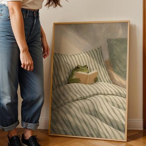 May include: A framed art print featuring a green frog reading a book in bed. The frog is positioned on a striped pillow and comforter, with a neutral-toned background. The frame is a light wood color.
