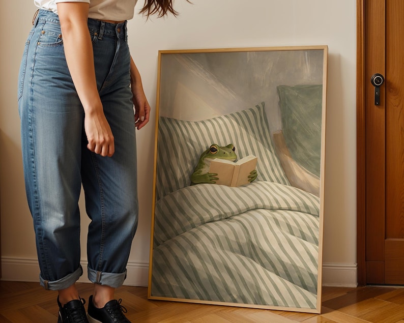 May include: A framed art print featuring a green frog reading a book in bed. The frog is positioned on a striped pillow and comforter, with a neutral-toned background. The frame is a light wood color.