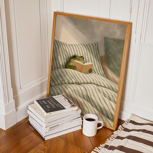 May include: A framed print depicts a green frog reading a book in bed, surrounded by green and white striped bedding. A stack of books, a white mug with dark liquid, and a brown and white striped rug are in the foreground.