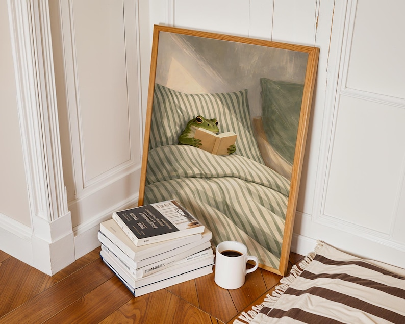 May include: A framed print depicts a green frog reading a book in bed, surrounded by green and white striped bedding. A stack of books, a white mug with dark liquid, and a brown and white striped rug are in the foreground.