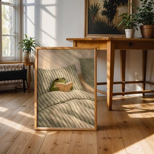 May include: A framed print featuring a whimsical illustration of a green frog reading a book in bed. The bedding has a striped pattern in shades of green and white. The frame is a light wood color, and the print is leaning against a wall in a room with wooden floors.