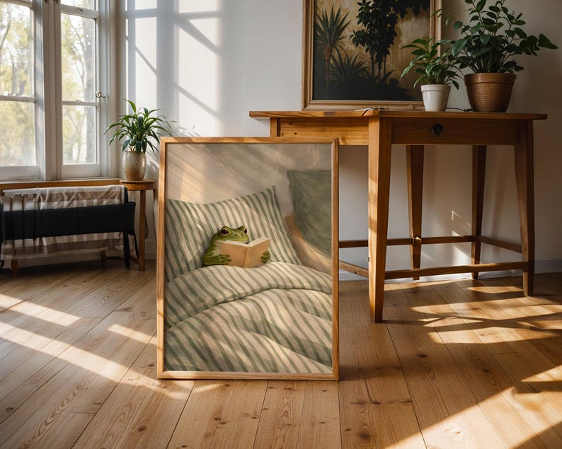 May include: A framed print featuring a whimsical illustration of a green frog reading a book in bed. The bedding has a striped pattern in shades of green and white. The frame is a light wood color, and the print is leaning against a wall in a room with wooden floors.