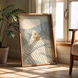 May include: A framed art print depicts a white duck reading a book in bed. The artwork features a light blue and white striped bedding design. The frame is made of wood, and the scene is set in a room with natural light.