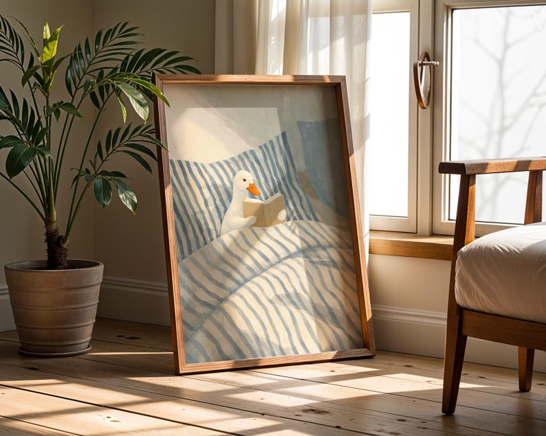 May include: A framed art print depicts a white duck reading a book in bed. The artwork features a light blue and white striped bedding design. The frame is made of wood, and the scene is set in a room with natural light.