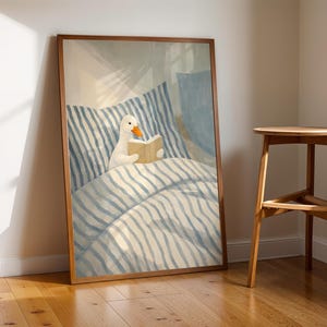 May include: A framed art print depicts a white duck reading a book in bed. The bedding has blue and white stripes. The artwork is in a wooden frame and leans against a wall. A wooden stool is next to the artwork.