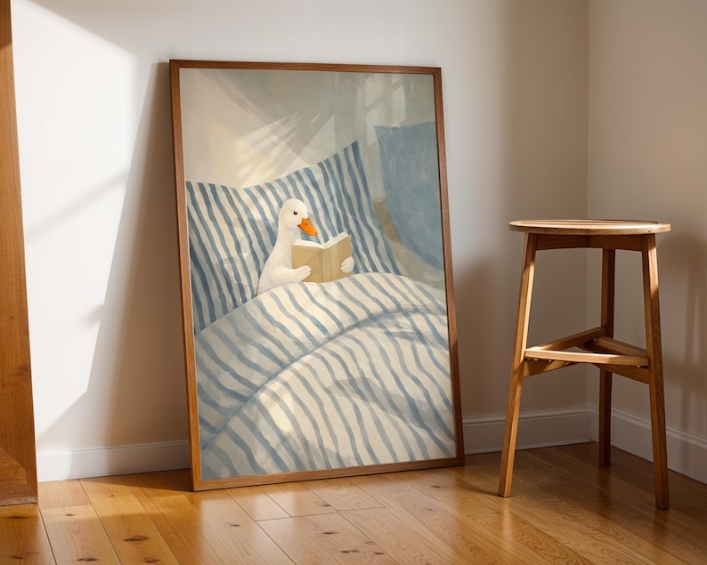 May include: A framed art print depicts a white duck reading a book in bed. The bedding has blue and white stripes. The artwork is in a wooden frame and leans against a wall. A wooden stool is next to the artwork.