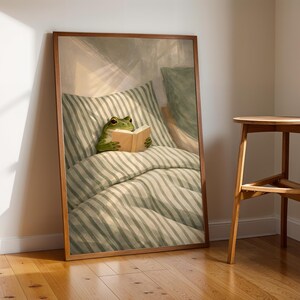 May include: A framed art print depicts a green frog reading a book in bed. The frog is nestled in a striped pillow and comforter. The artwork is in a wooden frame, leaning against a wall next to a wooden stool.
