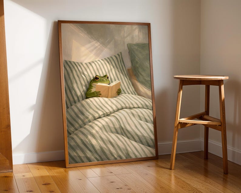 May include: A framed art print depicts a green frog reading a book in bed. The frog is nestled in a striped pillow and comforter. The artwork is in a wooden frame, leaning against a wall next to a wooden stool.