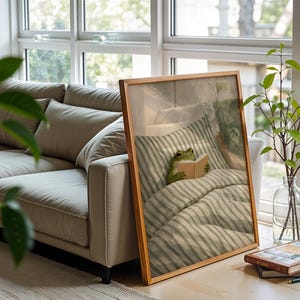 May include: A framed print featuring a green frog reading a book in bed. The artwork has a wooden frame and is propped against a window. A beige sofa and a vase with green branches are also visible.