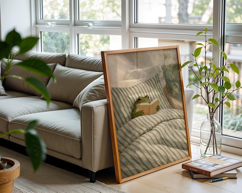 May include: A framed print featuring a green frog reading a book in bed. The artwork has a wooden frame and is propped against a window. A beige sofa and a vase with green branches are also visible.