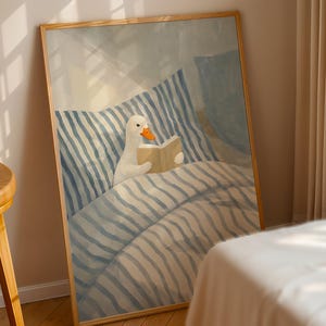 May include: A framed art print depicts a white goose reading a book in bed. The goose is nestled under a blue and white striped comforter. The artwork has a soft, pastel color palette and a wooden frame.