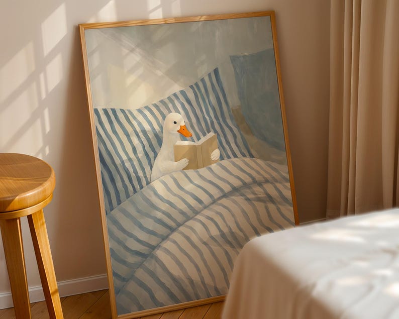 May include: A framed art print depicts a white goose reading a book in bed. The goose is nestled under a blue and white striped comforter. The artwork has a soft, pastel color palette and a wooden frame.
