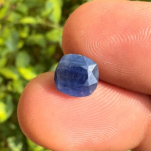 May include: A faceted, cushion-cut sapphire gemstone. The sapphire is a deep, rich blue color and has multiple facets that catch the light. The gemstone is held between two fingers, with a blurred green background.