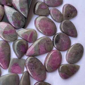 May include: A selection of polished gemstones in various shapes, such as ovals and teardrops. The stones display a combination of grey and pink colours, with a shiny surface. These gemstones are suitable for jewellery making or collecting.