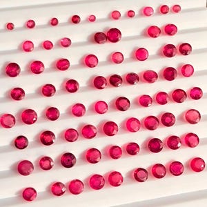 Pode incluir: Uma coleção de numerosas pedras preciosas redondas, facetadas e de um vibrante vermelho rosado, dispostas em filas numa bandeja de apresentação branca. Cada gema apresenta um corte brilhante com múltiplas facetas que refletem a luz.