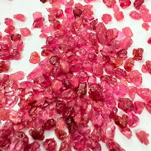 May include: A collection of small, rough-cut, ruby-red gemstones scattered on a white surface. The gemstones vary in size and shape, with a range of pink and red hues. The lighting highlights the facets and textures of the raw crystals.