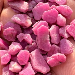 Puede incluir: Una colección de piedras preciosas de rubí en bruto y sin cortar. Las piedras son de un color rosa vibrante, con una textura rugosa y natural. Las piedras preciosas se sostienen en una mano.