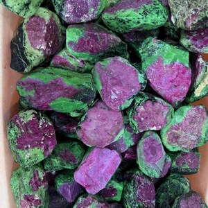 Peut inclure: Un ensemble de pierres brutes de zoisite et de rubis. Les pierres présentent un mélange de couleurs, avec des inclusions de rubis magenta vif sur une matrice de zoisite verte. Les pierres varient en taille et en forme, créant une esthétique naturelle et organique.