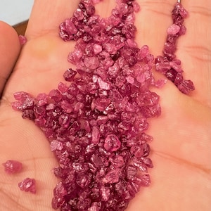Puede incluir: Primer plano de una mano que sostiene una colección de pequeñas piedras preciosas de color rosa intenso, talladas en bruto. Las gemas tienen forma irregular y un aspecto ligeramente translúcido. Las piedras están dispersas, creando un juego de color y luz.