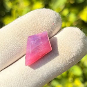 May include: A faceted gemstone with a gradient of pink and purple hues. The stone is held in a white glove, with a blurred green background. The gemstone's shape is a modified rhombus, showcasing its polished surfaces and color transitions.