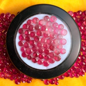 May include: A collection of small, round, faceted red gemstones. A large group of these loose stones is contained within a circular black display case with a white interior, while many more identical red gemstones are scattered around the base.