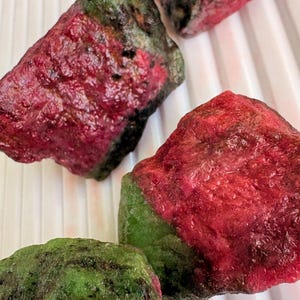 以下が含まれることがあります： いくつかの原石のクローズアップ。石は、鮮やかな赤と深い緑の色合いの色の混合を示しています。宝石は不規則な形をしており、質感のある表面で、自然に見えます。石は白い、溝のある表面に配置されています。