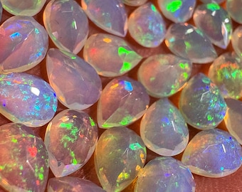May include: A collection of pear-shaped opals with a milky white base and iridescent flashes of color. The gemstones are faceted, catching and reflecting light with hues of green, blue, and orange. The opals are displayed against a warm, reddish background.