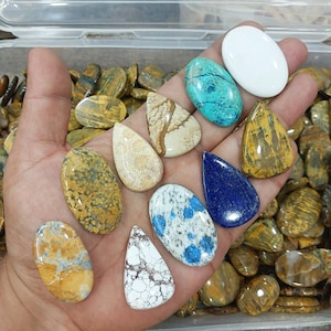 May include: A variety of polished gemstones in different shapes and colors are displayed in a hand. The stones include oval, teardrop, and irregular shapes, with colors like white, blue, yellow, and brown. Some stones have unique patterns and textures.