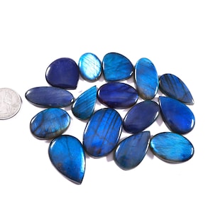 May include: A collection of polished, blue labradorite gemstones in various shapes, including oval, teardrop, and irregular forms. The stones display iridescent flashes of blue and are set against a white background. A US quarter is included for size reference.