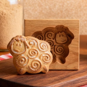 May include: A ceramic sheep-shaped cookie and wooden mold sit on a wooden cutting board with a red and white checkered cloth. A lit candle, pine cone, and glass jar with brown powder are also present. Two leaf-shaped cookies and an acorn are on the table.