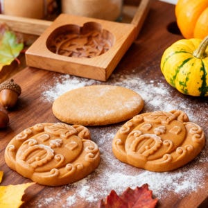 May include: A wooden pumpkin-shaped mould and two pumpkin-shaped biscuits sit on a wooden chopping board. The biscuits are surrounded by autumn leaves, acorns, and a small pumpkin. The biscuits are light brown and dusted with white powder.