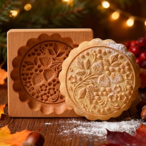 May include: A wooden cookie mold and a decorated cookie with a floral design. The mold is square with a scalloped edge, and the cookie has a detailed pattern of flowers and berries. The scene includes autumn leaves and a blurred background with lights.