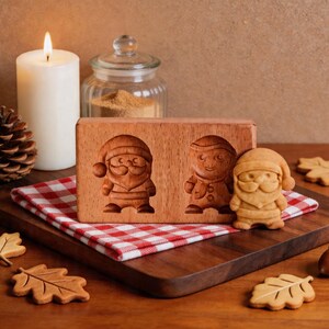 Könnte beinhalten: Holz-Keksform mit zwei Weihnachtsmann-Designs. Ein gebackener Keks in Form eines Weihnachtsmanns befindet sich neben der Form. Die Form und der Keks liegen auf einem Holzbrett mit blattförmigen Keksen.
