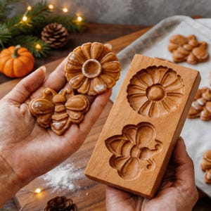 May include: A wooden mould featuring flower and bee designs, with baked biscuits in matching shapes. The mould is rectangular and made of light brown wood. The biscuits are golden brown and are on a white surface.
