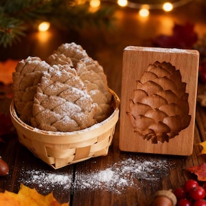 May include: A basket of pine cone-shaped cookies dusted with powdered sugar sits next to a wooden mold carved with a pine cone design. The cookies are light brown, and the basket is woven. Acorns and berries are scattered on the wooden surface.