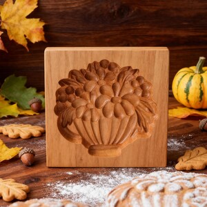 May include: A wooden cookie mold with a carved floral design. The mold features a detailed relief of flowers and leaves in a basket. The wood has a warm, natural tone. The image is set on a wooden surface with autumn leaves and a small pumpkin.