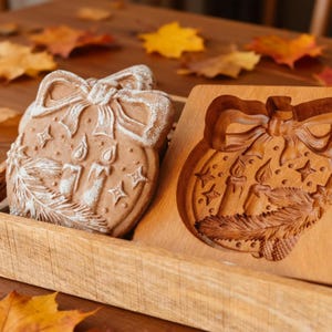 Pu&ograve; includere: Due biscotti a forma di ornamento natalizio con dettagli bianchi, accanto a uno stampo di legno. I biscotti presentano un fiocco, candele, stelle e un disegno di rametti. Lo stampo di legno ha lo stesso disegno intagliato.
