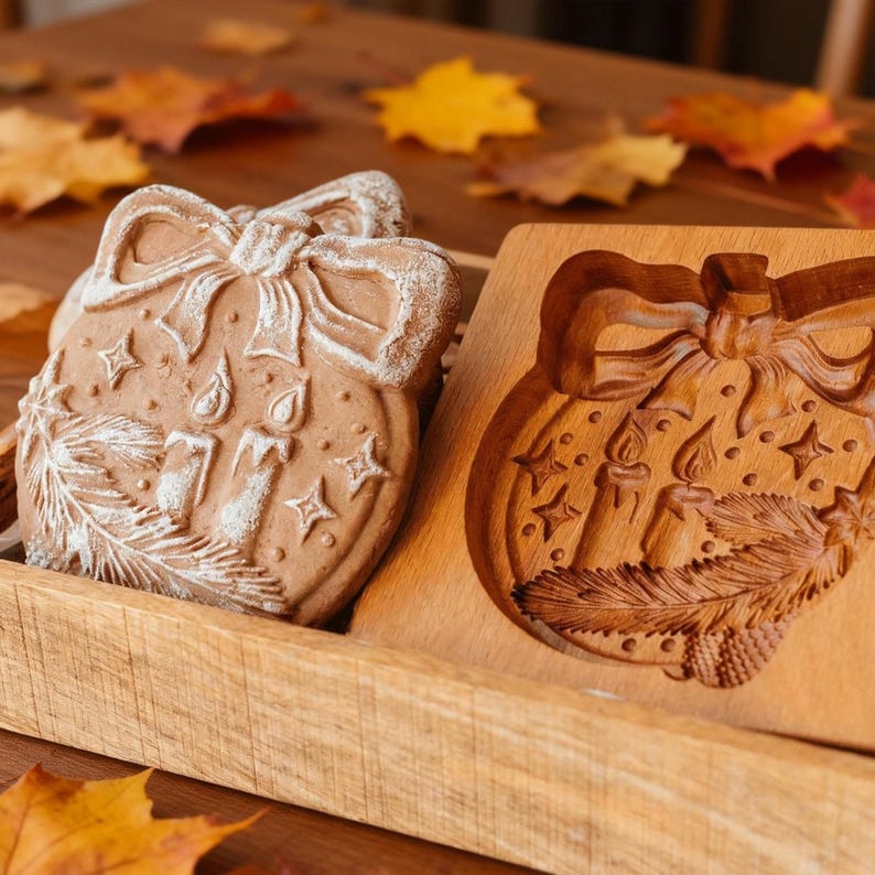 Pu&ograve; includere: Due biscotti a forma di ornamento natalizio con dettagli bianchi, accanto a uno stampo di legno. I biscotti presentano un fiocco, candele, stelle e un disegno di rametti. Lo stampo di legno ha lo stesso disegno intagliato.