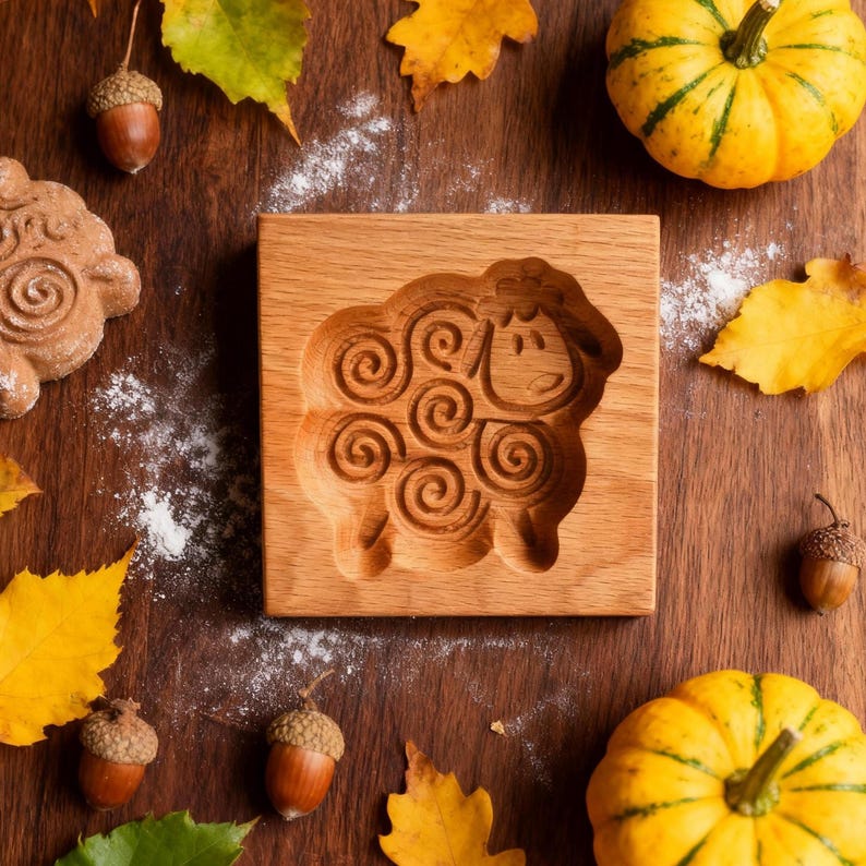May include: A wooden sheep-shaped cookie mold sits on a wooden surface, surrounded by autumn leaves, acorns, and small pumpkins. The mold features a detailed carving of a sheep with spiral wool patterns. The scene evokes a rustic, seasonal theme.